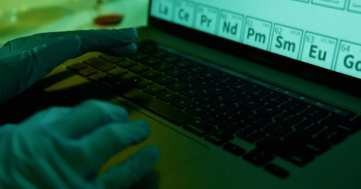 Un scientifique en gants saisit des données sur un ordinateur portable dans un laboratoire, concentré sur le tableau périodique.